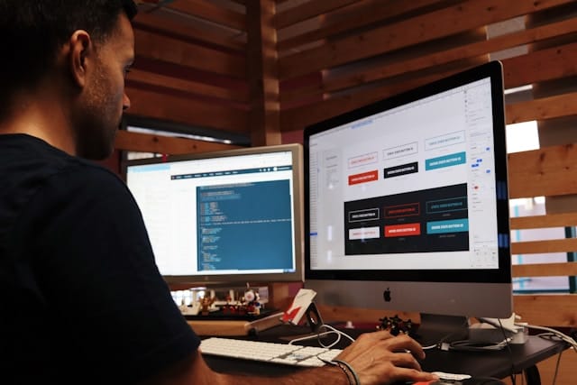 Developer building a website, with code on one monitor and a UI button-design mockup on an iMac